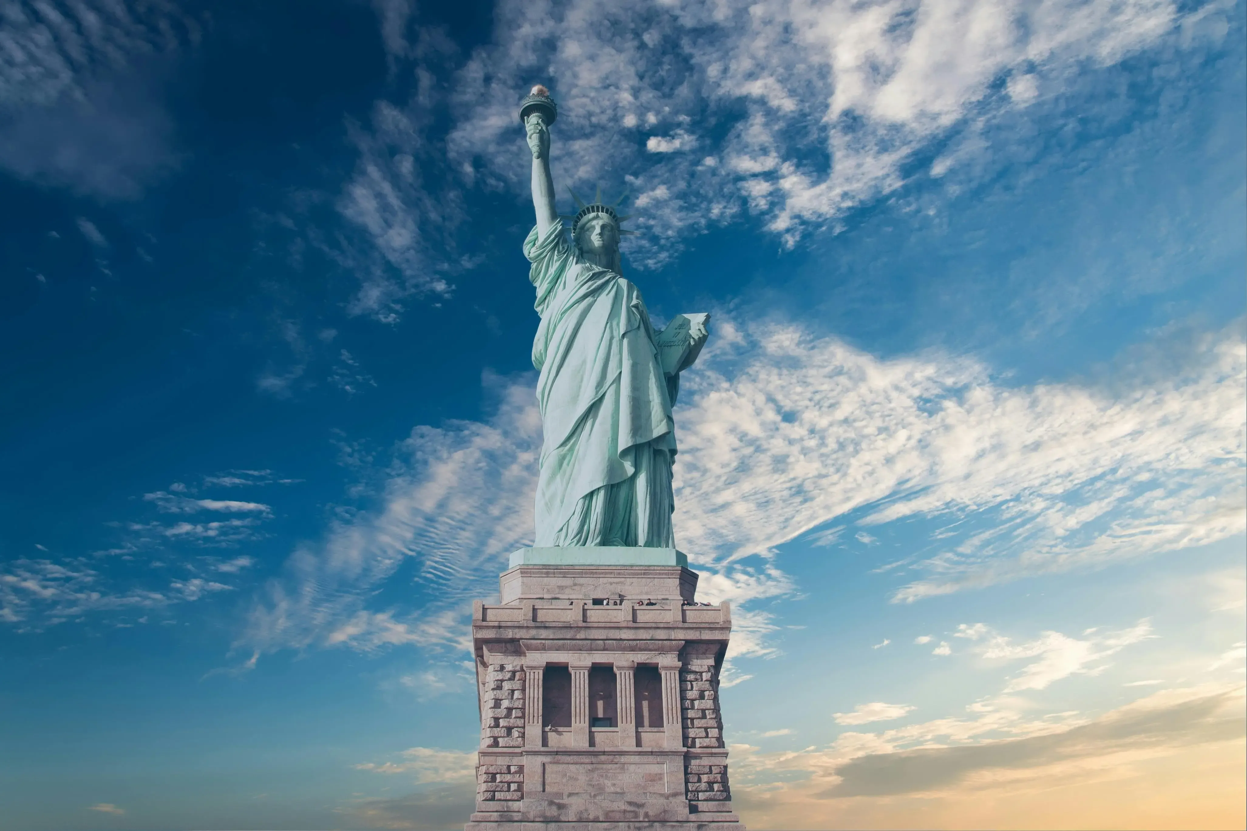 The Statue of Liberty standing tall against a blue sky with Ellis Island in the background, representing New York’s history.